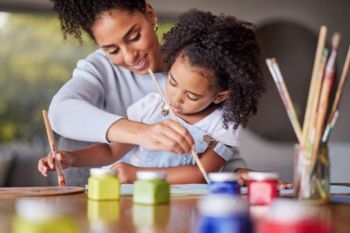 A mother and child painting together, with jars of colorful paint on the table.